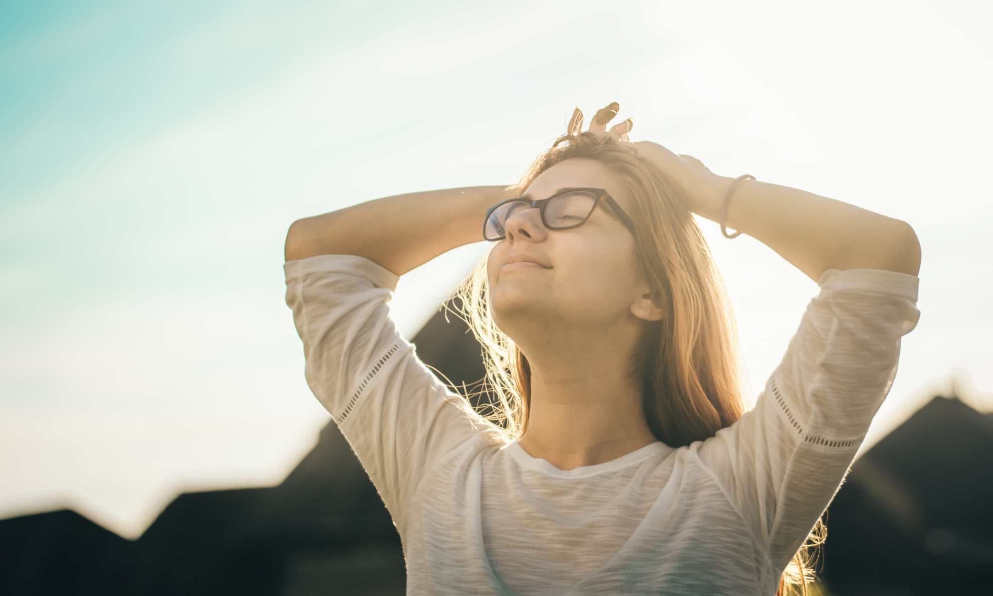Blonde Frau mit schwarzer Brille, genussvoll mit geschlossenen Augen, vor Bergsilhouette